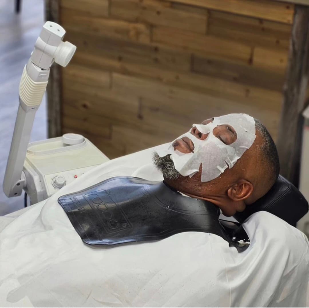 Man with a white sheet mask receives a facial treatment next to a professional steamer.